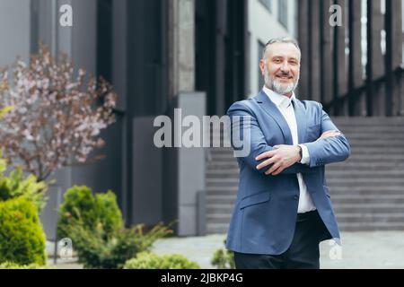 Uomo di successo capo di affari guardando la macchina fotografica con le braccia incrociate e sorridenti, uomo d'affari grigio-capelli in tuta di affari Foto Stock