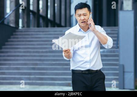 Frustrato e arrabbiato uomo d'affari asiatico che parla al telefono, tenendo una lettera e documenti da una banca fuori dell'ufficio Foto Stock