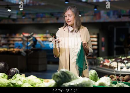 Senior bella donna shopping per le verdure nel negozio. Supermercato. Si alza, tiene il telefono nelle mani, tiene la rete alimentare, sceglie. Lui loo Foto Stock