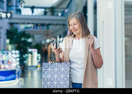 Ritratto di una bella donna anziana shopping nel centro commerciale. Lei usa il telefono, fa acquisti online, ha vinto un regalo, si rallegra. Contiene borse per la spesa in carta Foto Stock