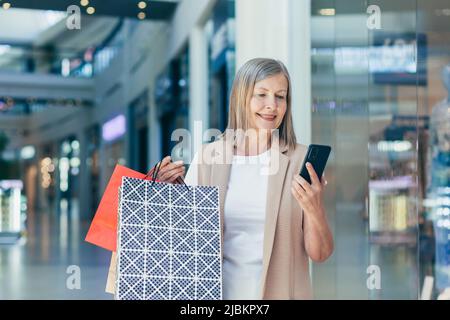 Donna anziana felice guarda lo schermo del telefono e si rallegra per lo sconto, un pensionato in un supermercato tiene nelle sue mani sacchetti colorati shopping Foto Stock