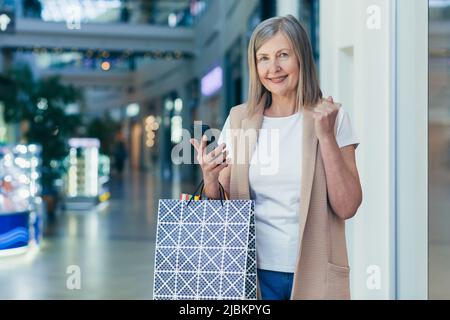 Ritratto di una bella donna anziana shopping nel centro commerciale. Lei usa il telefono, fa acquisti online, ha vinto un regalo, si rallegra. Contiene borse per la spesa in carta Foto Stock