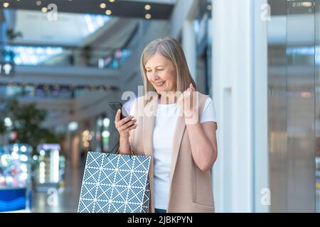 Donna anziana felice guarda lo schermo del telefono e si rallegra per lo sconto, un pensionato in un supermercato tiene nelle sue mani sacchetti colorati shopping Foto Stock