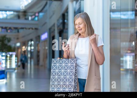 Ritratto di una bella donna anziana shopping nel centro commerciale. Lei usa il telefono, fa acquisti online, ha vinto un regalo, si rallegra. Contiene borse per la spesa in carta Foto Stock