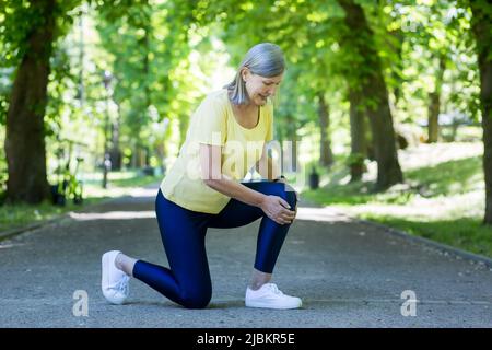 Trauma. Donna bella senior ferito il ginocchio. Tenendo le mani sulla gamba, si sedette. Entra per lo sport e corre nel parco. Foto Stock