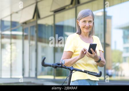 La donna anziana dai capelli grigi ama il telefono durante una passeggiata estiva con una bicicletta, sorridente pensionato Foto Stock