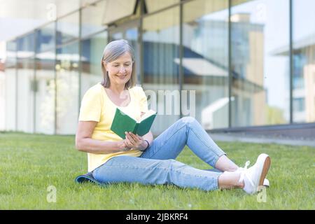 Ritratto di una bella donna anziana. Sedersi sull'erba, leggere un libro vicino ad un edificio moderno, riposa, ha un buon tempo Foto Stock