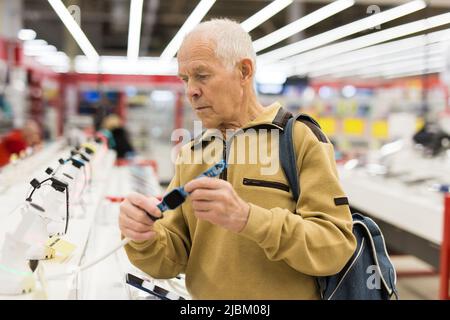 uomo anziano in grigio pensionato banco di esame con gadget elettronici e smart watch in showroom del negozio di merci digitali Foto Stock