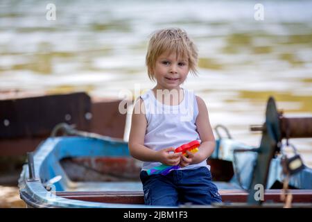 Bambino piccolo, ragazzo biondo, giocando con pop it antistress giocattolo nel parco in una barca in lago Foto Stock