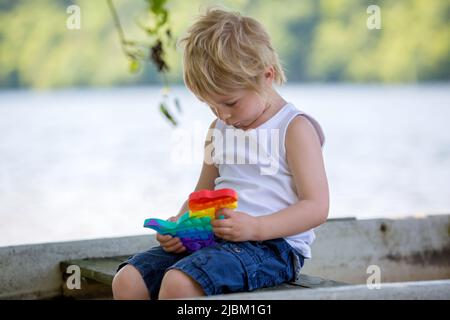 Bambino piccolo, ragazzo biondo, giocando con pop it antistress giocattolo nel parco in una barca in lago Foto Stock