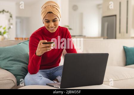 Giovane donna biraciale in foulard facendo shopping online attraverso la carta di credito a casa Foto Stock