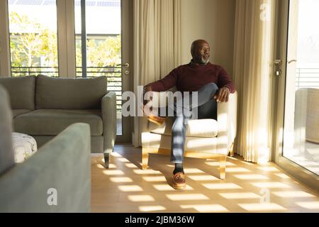 Un anziano afro-americano attento che guarda attraverso la finestra mentre si siede su una poltrona a casa Foto Stock