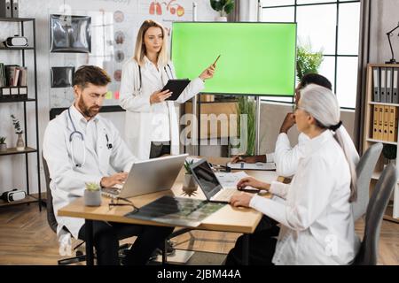 Medico caucasico femmina ponting sul monitor con schermo verde mock up mentre si fa la presentazione per i colleghi internazionali. Gruppo di esperti medici seduti alla scrivania e ascoltare briefing discorso. Foto Stock