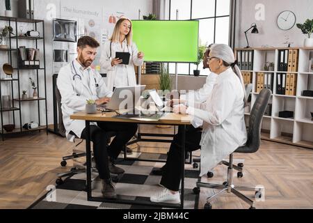 Medico caucasico femmina ponting sul monitor con schermo verde mock up mentre si fa la presentazione per i colleghi internazionali. Gruppo di esperti medici seduti alla scrivania e ascoltare briefing discorso. Foto Stock