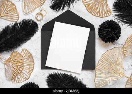 Carta bianca e busta nera sul tavolo in marmo vicino a piume nere e foglie dorate con vista dall'alto degli anelli da matrimonio. Biglietto di auguri o invito mockup. E Foto Stock
