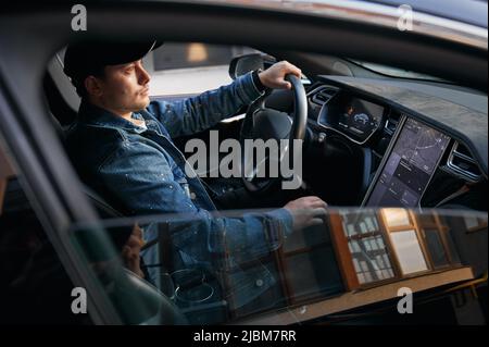 Vista attraverso il finestrino laterale aperto dell'auto. Uomo in berretto seduto in automobile d'elite, mettendo una mano sul volante e d'altra parte inserendo l'indirizzo di destinazione sul grande touch screen. Foto Stock