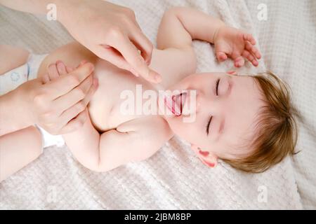 Le mani della madre stanno spazzolando i denti di un bambino felice del toddler su un letto domestico. La mamma spazzola la bocca del bambino con uno spazzolino. Bambino di un anno Foto Stock