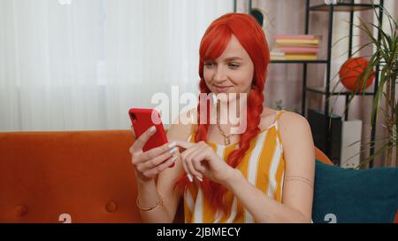 Donna allegra seduta sul divano usa il sorriso del telefono cellulare in casa moderna appartamento. Happy ginger girl texting Condividi i contenuti dei messaggi sulle applicazioni social media per smartphone online, guardando film relax Foto Stock