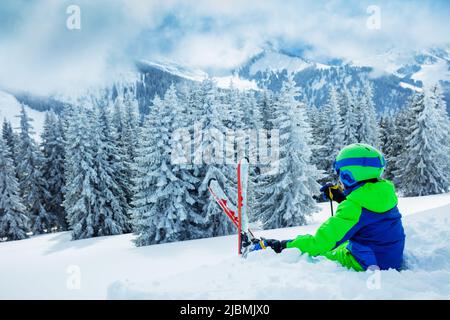 Il bambino si siede nella neve sopra la foresta innevata di abete dopo la nevicata pesante Foto Stock