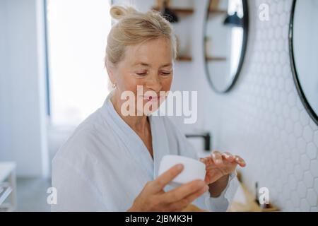 Bella donna anziana in accappatoio applicazione crema naturale in bagno, cura della pelle e mattina concetto di routine. Foto Stock