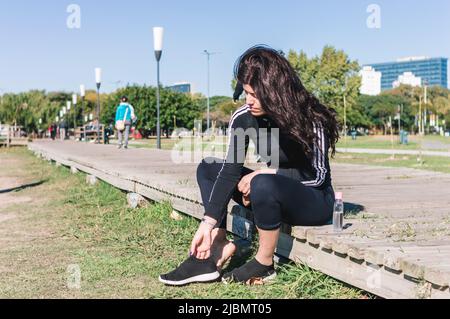 bella latina caucasica giovane donna indossando abbigliamento sportivo nero, seduta all'aperto mettendo le sue scarpe per iniziare l'allenamento e il jogging. Foto Stock