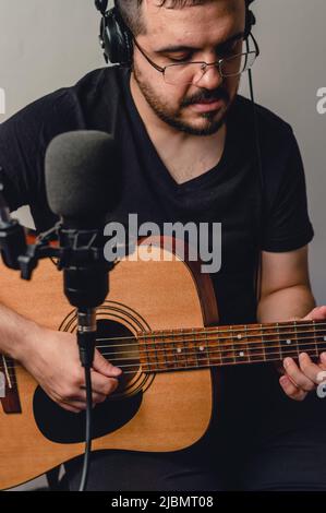 uomo venezuelano latino caucasico, con occhiali, cuffie e vestiti neri, seduto in studio di musica, che registra una canzone che suona chitarra acustica e canta Foto Stock