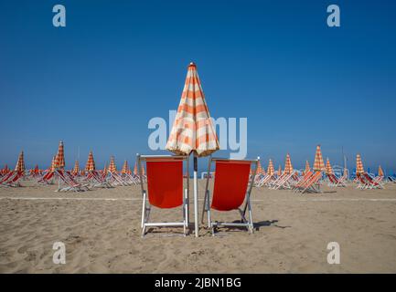 Una vista di sdraio rosse sulla spiaggia con ombrelloni arancioni e bianchi in una giornata di sole con un cielo azzurro. Foto Stock
