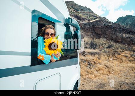 Moderno stile di vita da viaggio hippy. Felice giovane donna fuori dal camper van finestra godere di viaggio destinazione e tenendo grande girasole. Concetto di affitto veh. rv Foto Stock