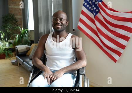 Ritratto di uomo afroamericano sorridente alla macchina fotografica mentre si siede in sedia a rotelle contro la bandiera americana sulla parete nella stanza Foto Stock