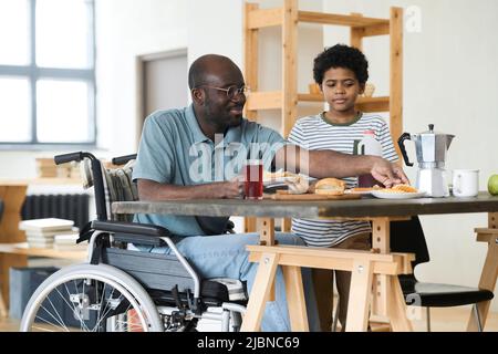 Singolo padre africano seduto in sedia a rotelle al tavolo e fare colazione con suo figlio al mattino a casa Foto Stock