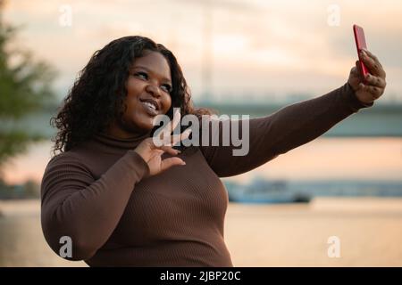 Sorridente e gioiosa bella afroamericana donna in abito elegante prendendo selfie da smartphone. Vacanze estive vicino al mare Foto Stock