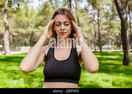 Giovane bella donna che indossa abiti sportivi nel parco cittadino, all'aperto tenendo la testa con le mani cercando di ricordare qualcosa o avere mal di testa. SUF Foto Stock