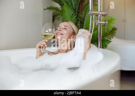 Bella giovane donna felice godendo bolle schiuma e vino bianco nella vasca idromassaggio. Foto Stock