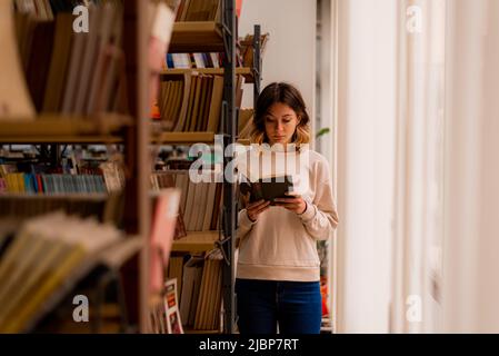 College girl leggendo un libro mentre si trova accanto alla finestra nella biblioteca Foto Stock