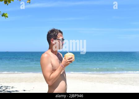 Giovane uomo che beve cocco sulla spiaggia di sabbia Foto Stock