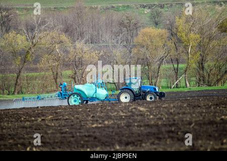 Trattore blu che spruzza insetticida su terreno coltivato campo. Attrezzature agronomiche agricole in funzione in una giornata di primavera. Paesaggio rurale Foto Stock