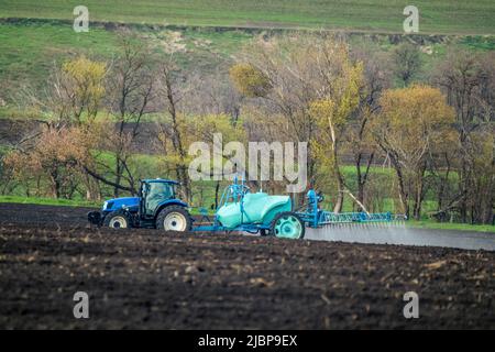 Trattore blu che spruzza insetticida su terreno coltivato campo. Macchine agricole contadine che lavorano in una giornata di primavera. Paesaggio rurale Foto Stock