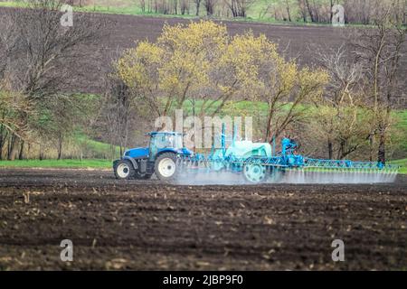 Trattore che spruzza insetticida su campo di terra coltivato. Attrezzature agronomiche agricole in attività nei terreni agricoli primaverili. Paesaggio rurale Foto Stock