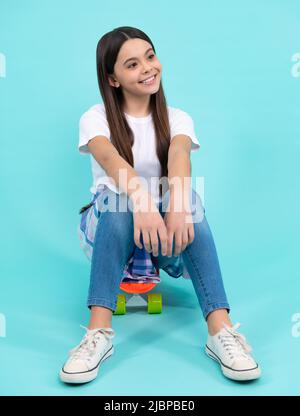 Buon vogue adolescente. Ragazza giovane alla moda e sicura, con t-shirt e jeans alla moda, si siede sullo skateboard, posando su sfondo blu. Moda Foto Stock