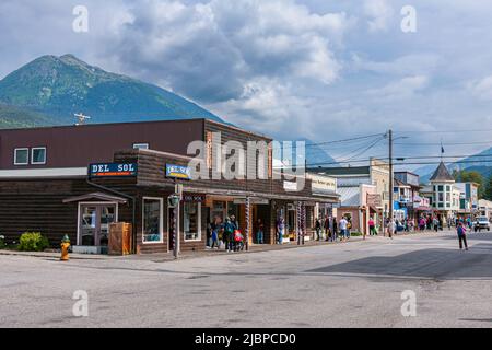 Skagway, Alaska, USA - 20 luglio 2011: Del Sol e altri negozi al dettaglio su Broadway sotto il paesaggio blu e verde montagna sul retro. Persone vestiti e. Foto Stock