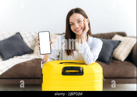 Bella ragazza caucasica con occhi blu in abiti eleganti, si siede a casa nel soggiorno vicino a una grande valigia gialla, mostra uno smartphone con uno schermo bianco mockup, guarda la fotocamera, sorride Foto Stock