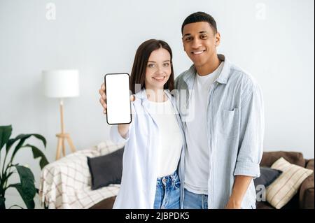 Tecnologia wireless. Felice coppia multirazziale moderno stand in salotto, mostrando uno smartphone con uno schermo bianco bianco bianco mockup per la pubblicità, guardano la fotocamera con eccitato, sorridente Foto Stock