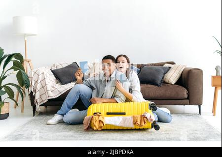 Felice famiglia coppia multirazziale, ragazzo razza mista e ragazza caucasica, sedersi sul pavimento a casa nel soggiorno vicino a una grande valigia gialla, tenere passaporti, preparare per una vacanza a lungo atteso, sorriso Foto Stock