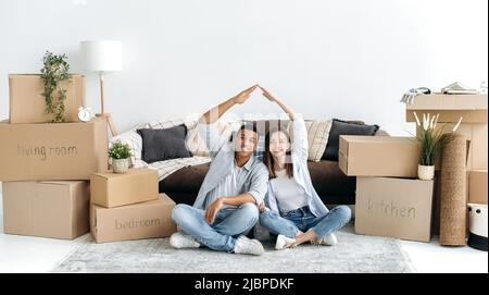 Foto panoramica di gioiosa giovane coppia innamorata seduta sul pavimento nel soggiorno della loro nuova casa tra scatole di cartone, facendo un tetto immaginario con le mani, guardando la macchina fotografica, sorridendo Foto Stock