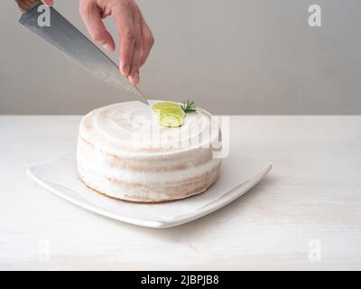 La mano di un uomo taglia una torta di compleanno Vegan con mandorle, cocco e limone base con un coltello grande su un tavolo bianco e uno sfondo bianco Foto Stock