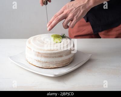 La mano di un uomo taglia una torta di compleanno Vegan con mandorle, cocco e limone base con un coltello grande su un tavolo bianco e uno sfondo bianco Foto Stock