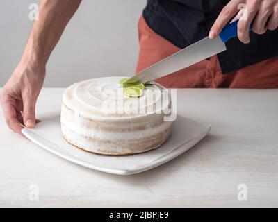 La mano di un uomo taglia una torta di compleanno Vegan con mandorle, cocco e limone base con un coltello grande su un tavolo bianco e uno sfondo bianco Foto Stock