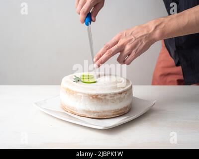 La mano di un uomo taglia una torta di compleanno Vegan con mandorle, cocco e limone base con un coltello grande su un tavolo bianco e uno sfondo bianco Foto Stock