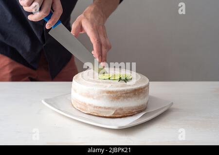 La mano di un uomo taglia una torta di compleanno Vegan con mandorle, cocco e limone base con un coltello grande su un tavolo bianco e uno sfondo bianco Foto Stock