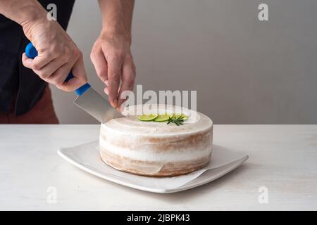 La mano di un uomo taglia una torta di compleanno Vegan con mandorle, cocco e limone base con un coltello grande su un tavolo bianco e uno sfondo bianco Foto Stock